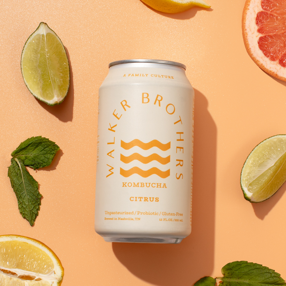 Can of Walker Brothers Kombucha Citrus kombucha placed on a peach-colored background, surrounded by sliced citrus fruits (lime, lemon, grapefruit) and mint leaves. The can features an abstract logo with wavy lines in yellow and text indicating the beverage is unpasteurized, probiotic, and gluten-free.