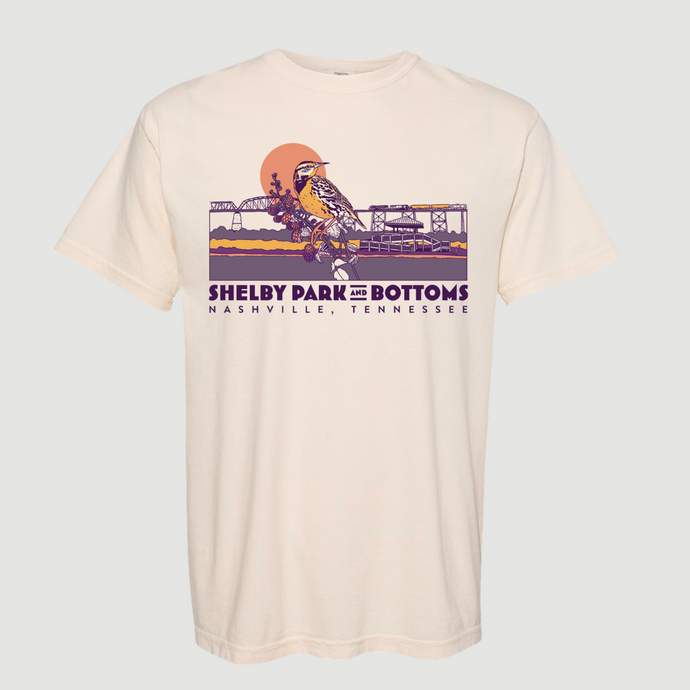 Cream colored t-shirt with Shelby Park and Bottoms Nashville, Tennessee in purple. Above the text there is an illustration of a bird on a branch in front a landscape drawing that features a train and grass and a sun. The primary colors of the illustration are shaedes of purple, coral and yellow.