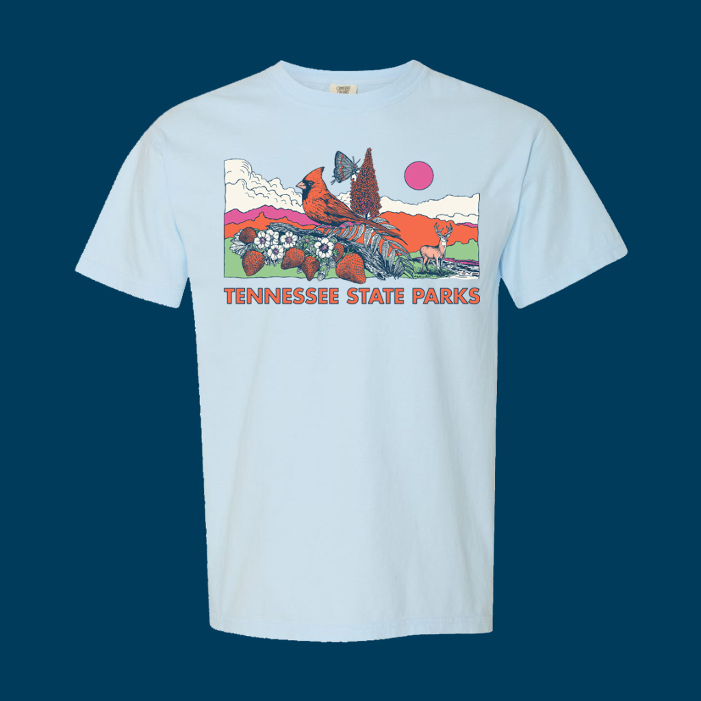 Light blue T-shirt with a colorful graphic showing a male cardinal, butterfly, white-tailed deer, sumac, strawberries, and blossoms against a scenic background of clouds, mountains and a pink sun. Below the illustration, bold orange text reads 'Tennessee State Parks.'